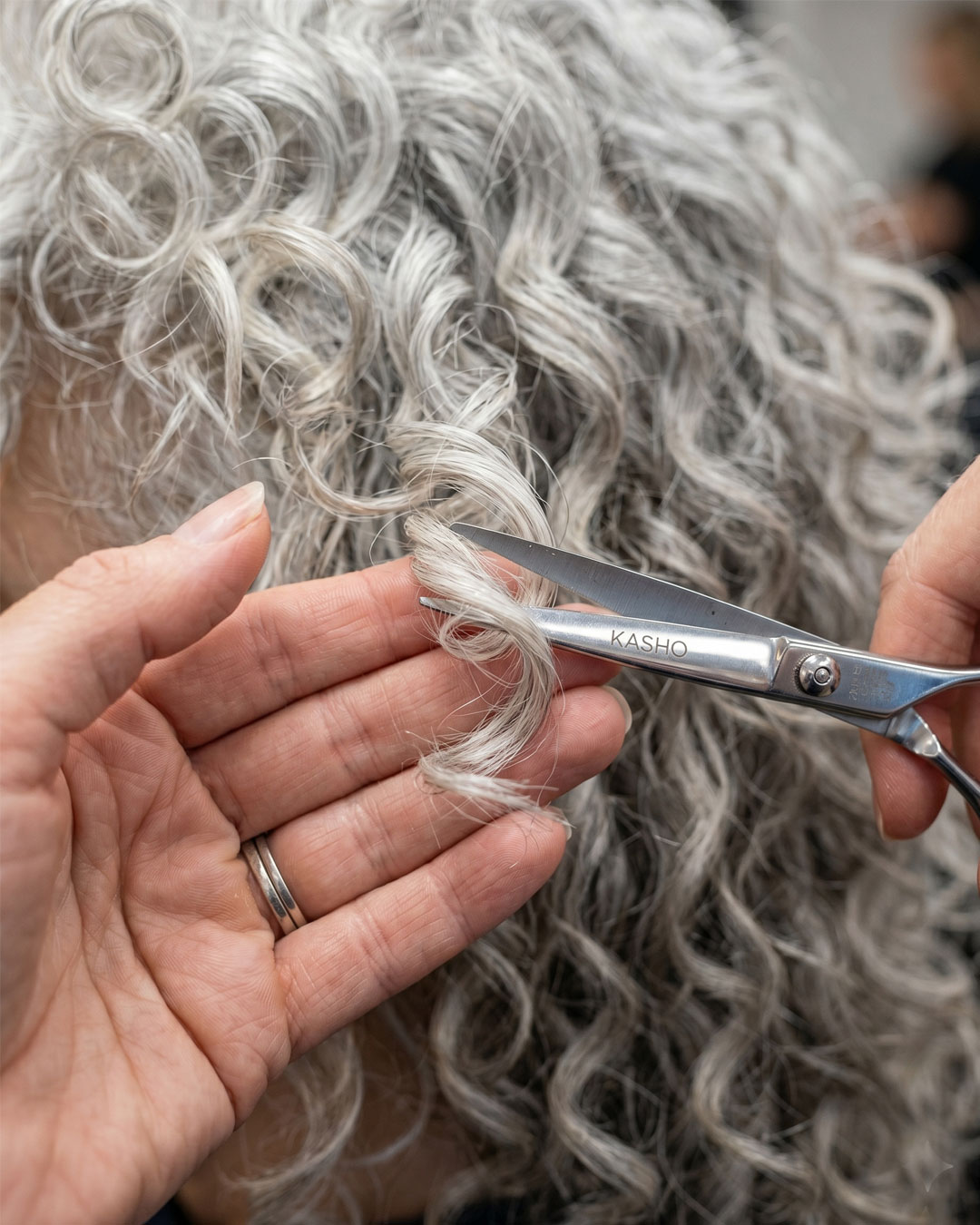 Stylist using scissors to dry-cut individual silver curls on a mature woman.